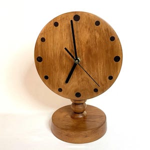 May include: A wooden desk clock with a round face and a pedestal base. The clock face is light brown with black dots marking the hours and black hands. The base is a darker brown. The clock is a decorative item.