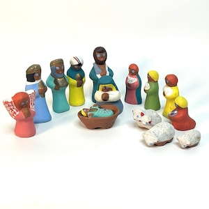 May include: A colorful ceramic nativity set featuring figures in various colors, including blue, yellow, and red. The set includes figures of the Holy Family, angels, and sheep. The figures are arranged on a white surface.