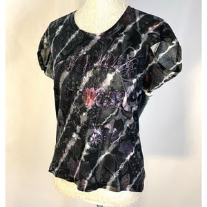 May include: Black, short-sleeved t-shirt with a round neckline. The shirt features a tie-dye effect with white, gray, and black stripes. Intricate floral and abstract patterns in shades of pink, purple, and gray are printed across the front.