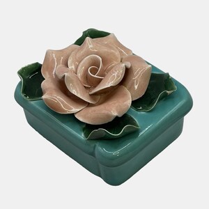 May include: A teal ceramic trinket box with a large, detailed, peach-coloured rose on the lid. The rose is surrounded by green leaves. The box has a rectangular shape with rounded edges.