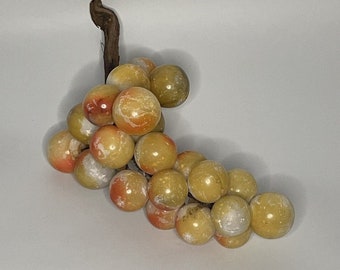 May include: A cluster of faux grapes with a wooden stem. The grapes are a mix of yellow, orange, and white, with a marble-like texture. The stem is a dark brown color. The grapes are arranged in a natural, cascading shape.