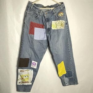 May include: Light blue denim jeans with various fabric patches. Patches include red polka dots, blue gingham, floral print, a brown square, a purple jellyfish, a SpongeBob SquarePants graphic, and yellow fabric. The jeans are hanging on a white hanger.