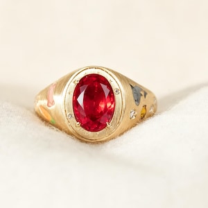May include: A gold signet ring featuring a large, oval-cut vibrant red gemstone at the center. The band is decorated with small, colorful enamel animal motifs and tiny embedded clear stones, set against a brushed, matte gold metal finish.
