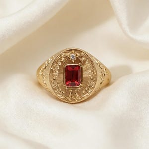 Puede incluir: Un anillo dorado con una piedra preciosa rectangular de color rojo intenso en el centro. El anillo presenta intrincados detalles grabados, incluido un diseño circular con motivos florales y de hojas. Una pequeña piedra transparente está engastada encima de la piedra roja.