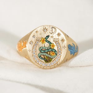 May include: A gold signet ring featuring a central horseshoe motif encrusted with small clear stones. Inside the horseshoe is a green snake entwined with yellow flowers. The ring surface includes engraved stars, a crescent moon, and the letter D.