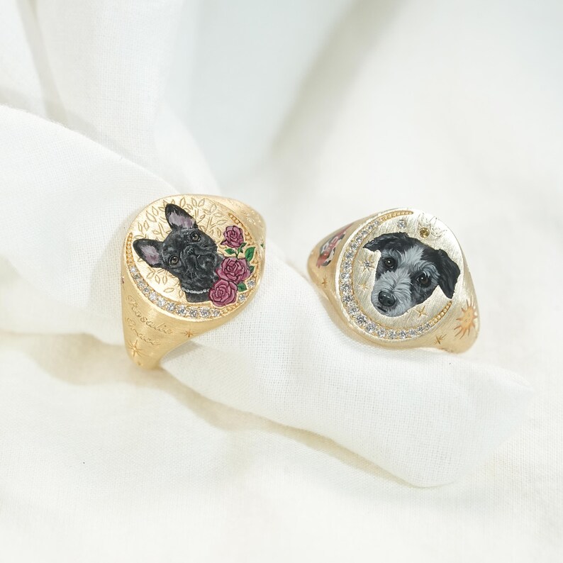 May include: Two gold signet rings with detailed portraits of dogs. One ring features a black French Bulldog with pink roses, the other a black and white terrier-mix. Both rings have a diamond-studded border and engraved details.