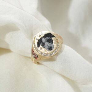 May include: Gold signet ring featuring a hand-painted portrait of a dog with black and white fur. The ring has a halo of small, clear stones around the dog portrait and is decorated with stars and a red and black emblem.