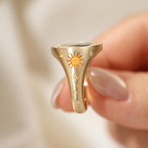 May include: A gold-colored ring with a sun and star design. The ring features a detailed sunburst design with a face, and several small stars with diamond accents. The ring is held up by a person's hand.
