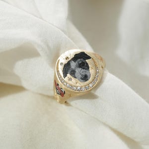 May include: A gold signet ring featuring a painted portrait of a black and white dog. The ring is adorned with small diamonds and star accents around the dog's portrait. A red heart with text is visible on the side of the ring.