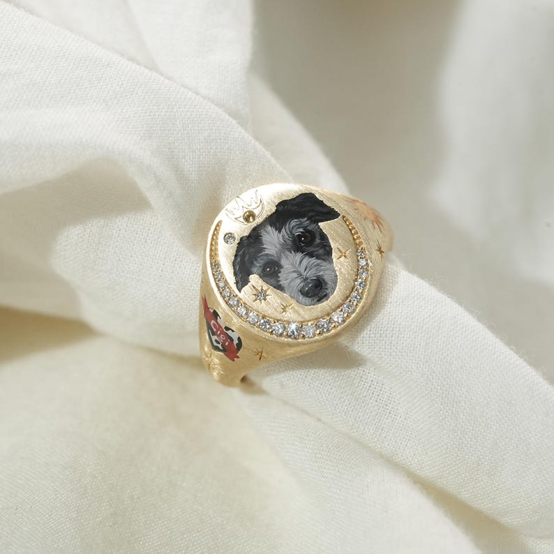 May include: A gold signet ring featuring a painted portrait of a black and white dog. The ring is adorned with small diamonds and star accents around the dog's portrait. A red heart with text is visible on the side of the ring.