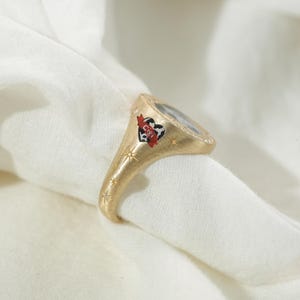 May include: Gold-toned ring with a heart-shaped emblem featuring the letters "CIGI" and a red banner. The ring has a textured finish and is adorned with small starburst designs along the band. The ring is set with small gemstones.