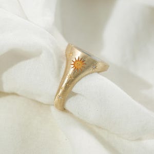 May include: Gold-toned ring featuring a sun design with a smiling face and star accents. The ring has a brushed finish and small, sparkling embellishments. The ring is set against a white fabric background.