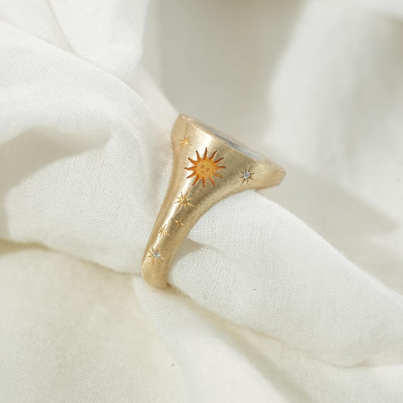 May include: Gold-toned ring featuring a sun design with a smiling face and star accents. The ring has a brushed finish and small, sparkling embellishments. The ring is set against a white fabric background.