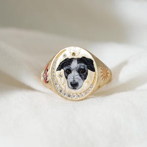 May include: Gold signet ring featuring a hand-painted dog portrait with black and white fur. The ring has a diamond-studded oval face, a crown, and sun and star details. The ring is a unique piece of jewelry.