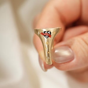 May include: A gold-colored signet ring with a heart-shaped emblem. The emblem features a black and white cow-print heart with the name "Cigi" on a red banner. The ring is adorned with starburst designs.