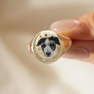 May include: A gold signet ring featuring a painted portrait of a dog with black and white fur. The ring has a crown, moon, and sun design. The ring is encrusted with small, clear gemstones.