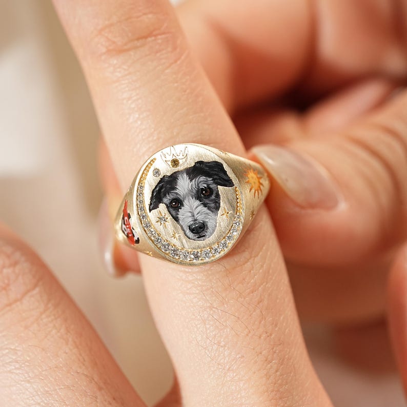 May include: Gold signet ring featuring a detailed portrait of a dog with black and white fur. The ring has a circular face with a crown and star accents, and is embellished with small, clear gemstones.