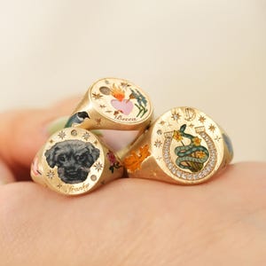 May include: Gold signet rings with detailed illustrations. One ring features a dog portrait with the name "Franky", another has a heart and floral design with the name "Becca", and the third ring has a snake and horseshoe design.