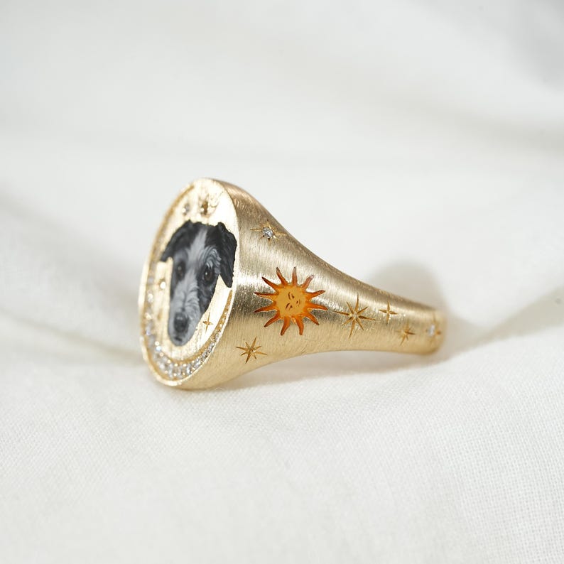 May include: A gold signet ring featuring a black and white dog portrait within a diamond-studded oval. The ring has a brushed finish, with a sunburst and star accents. A unique piece of jewelry.
