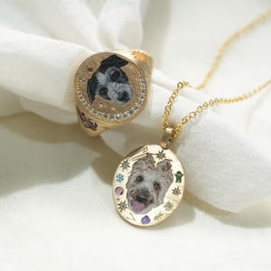 May include: A gold ring and pendant necklace featuring custom portraits of dogs. The ring has a round face with a black and white dog portrait, surrounded by small diamonds. The pendant has an oval face with a dog portrait and is suspended from a delicate gold chain.