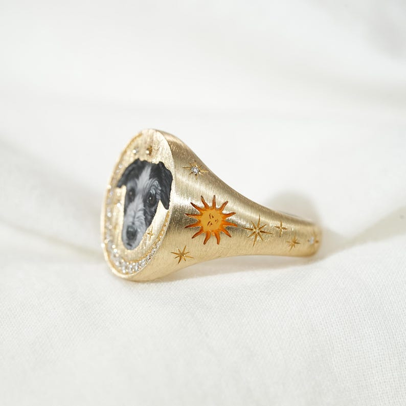 May include: Gold signet ring featuring a black and white dog portrait. The ring has a textured finish, a sun design, and star accents. The ring is adorned with small, clear gemstones around the dog portrait.