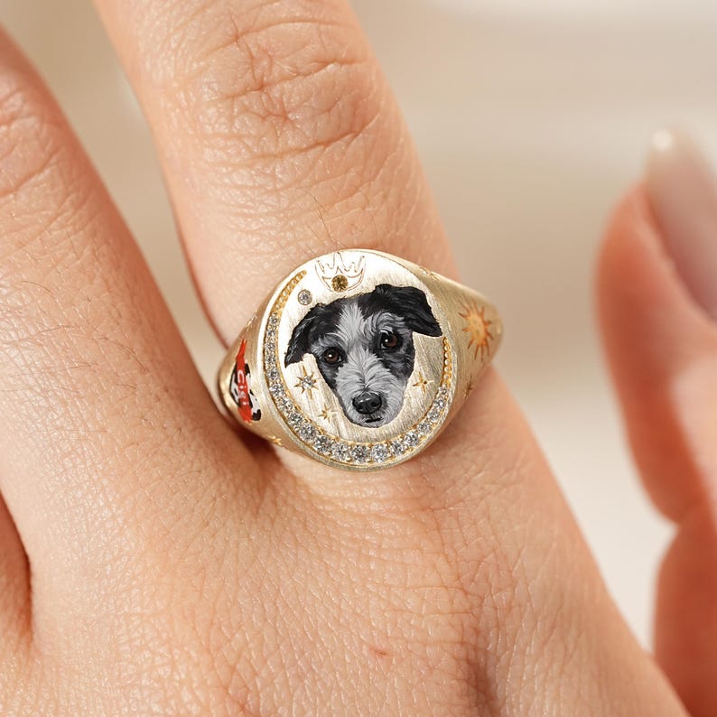 May include: A gold-colored signet ring featuring a hand-painted portrait of a dog with black and white fur. The ring is adorned with small diamonds, stars, and a sun design. The ring is on a finger.