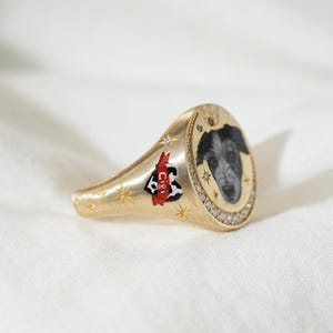 May include: Gold signet ring featuring a black and white dog portrait. The ring has a diamond-studded border and a heart-shaped design with the name "Cigi" on the side. The ring is decorated with star and diamond accents.