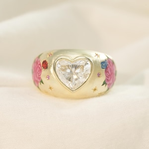May include: A gold ring with a heart-shaped diamond center. The ring is decorated with pink roses, blue flowers, and small gold stars. A unique piece of jewelry.