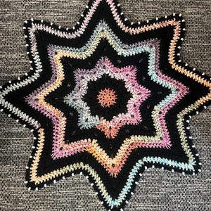 May include: A star-shaped crochet doily featuring a central orange circle surrounded by concentric layers of black yarn alternating with variegated pastel yarn in shades of pink, blue, and yellow, finished with a white-stitched black border.