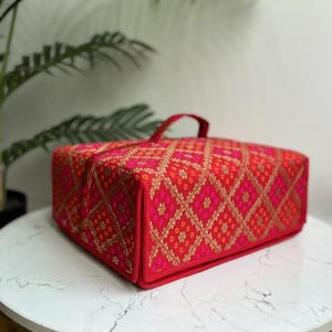 May include: A rectangular, red fabric bag with a handle, featuring a detailed pattern of gold and pink floral and diamond shapes. The bag is sitting on a white marble surface.