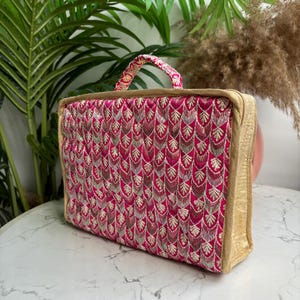 May include: A rectangular, decorative bag with a handle. The bag features a vibrant pink fabric with an embroidered pattern in gold and brown. The edges and handle are trimmed with gold fabric.