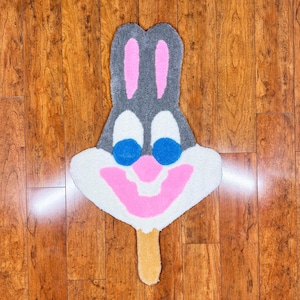May include: A gray and white rug shaped like a cartoon rabbit with blue eyes and a pink mouth. The rug has a brown stick-like shape at the bottom.