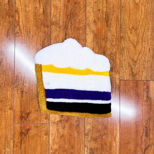 May include: A white, yellow, purple, and black layered cake rug. The rug is shaped like a slice of cake and has a fluffy texture.
