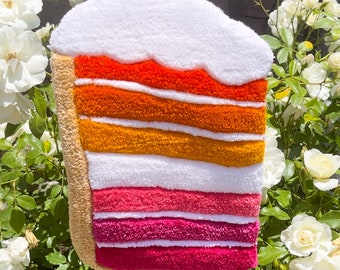 Op de afbeelding: Een kleurrijke, gelaagde cake gemaakt van vilt, met een witte frosting bovenop. De cake is in levendige oranje, gele, roze en rode tinten.