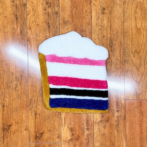 May include: A colorful striped rug shaped like a slice of cake with white frosting. The rug has a gold border and is made of soft, fluffy material.