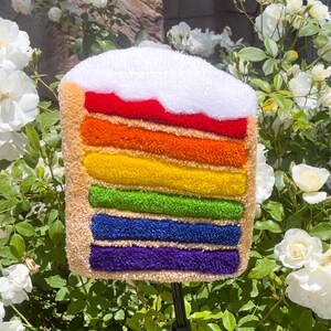May include: A rainbow layered cake with white frosting, made of yarn and displayed against a backdrop of white roses.