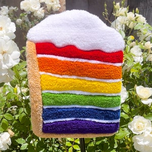 May include: A rainbow layered cake made of plush fabric. The cake is decorated with white frosting and has a brown base.