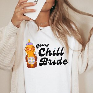 Könnte beinhalten: Weißes T-Shirt mit einem Grafikdruck eines Comic-Honigbären, der einen Schleier und eine Sonnenbrille trägt. Der Text "Bear-y Chill Bride" ist in Schwarz gedruckt.