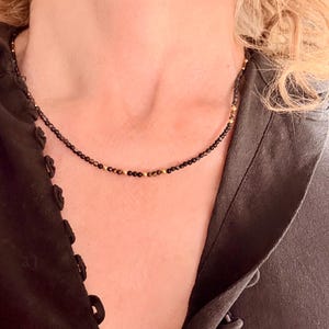 May include: A fine black beaded necklace with gold-coloured detailing. The choker-style necklace is made up of small, round beads. The necklace is set against a dark background, which accentuates the contrast of the colours.