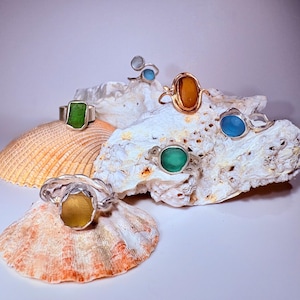 Könnte beinhalten: Eine Sammlung von sechs Ringen mit poliertem Meerglas in Grün-, Blau- und Bernsteintönen, gefasst in silberfarbene Metallringe. Die Ringe sind auf strukturierten weißen und orangefarbenen Muscheln arrangiert und zeigen verschiedene Formen.