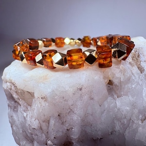May include: A beaded bracelet featuring amber-colored, cube-shaped beads interspersed with gold-colored geometric beads. The bracelet is displayed on a white, textured surface, showcasing its warm tones and intricate design. The bracelet is a piece of jewelry.