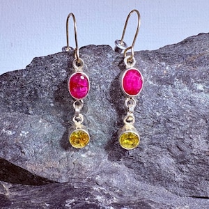 May include: A pair of silver dangle earrings. Each earring has a fuchsia oval gemstone at the top and a yellow-green oval gemstone at the bottom. The earrings are displayed on a dark grey, textured surface.