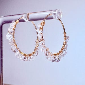 Peut inclure: Boucles d'oreilles créoles dorées avec des accents de cristal transparent. Les boucles d'oreilles sont faites de fil enroulé autour des cristaux.