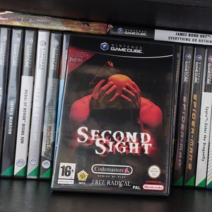 May include: A collection of video game cartridges for the Nintendo GameCube console. The cartridges are arranged vertically on a shelf. The front of the cartridge for the game "Second Sight" is visible. The game is rated 16+ and features a red hand reaching towards a glowing orb. The text "Second Sight" is in white letters on a black background.