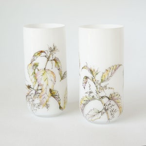 Op de afbeelding: Twee witte glazen bekers met een bloemenmotief. Het ontwerp bevat delicate gele, groene en bruine bladeren en bloemen.