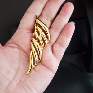 May include: A gold-colored, stylized wing-shaped brooch. The brooch features a series of curved, parallel lines, creating a layered effect. The design is smooth and polished, reflecting light. The brooch is held in a hand against a dark background.