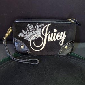 May include: Black clutch with the word "Juicy" in white script, and a crown design. The clutch has a wrist strap and a zipper closure. The corners are trimmed with black leather and gold rivets.