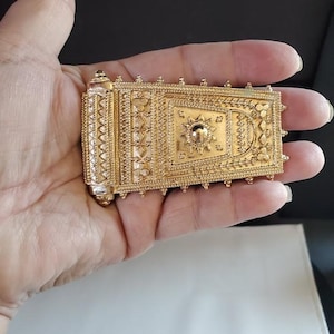 May include: A rectangular gold-toned metal pendant featuring intricate embossed patterns, including a central sunburst motif, geometric shapes, and small beaded edges. The piece has a cylindrical hinge-like element on one side.