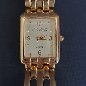 May include: A gold-toned rectangular wristwatch with a cream-colored face. The watch face displays the numbers 12, 3, 6, and 9, with the words "JOAN RIVERS CLASSICS" and "QUARTZ" printed on it. The watch band is also gold-toned.