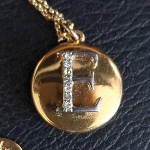 May include: A gold-toned pendant necklace featuring a circular charm with the letter "E". The letter is silver-toned and embellished with small, clear crystals. The pendant hangs from a delicate gold chain, creating a classic and personalized accessory.